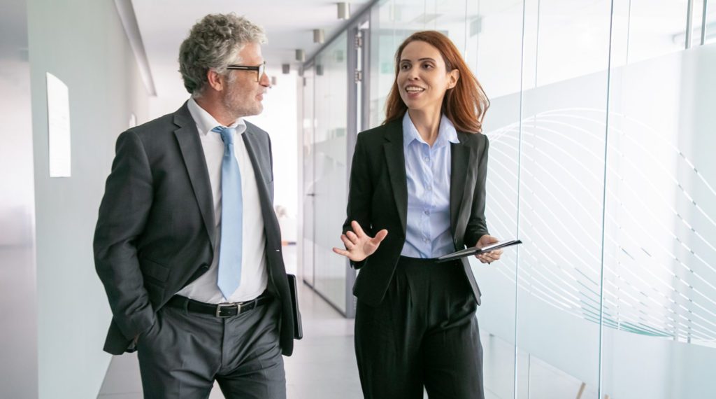 Successful businesspeople walking through office corridor and talking. Professional senior executive manager in eyeglasses listening beautiful content businesswoman. Business and management concept