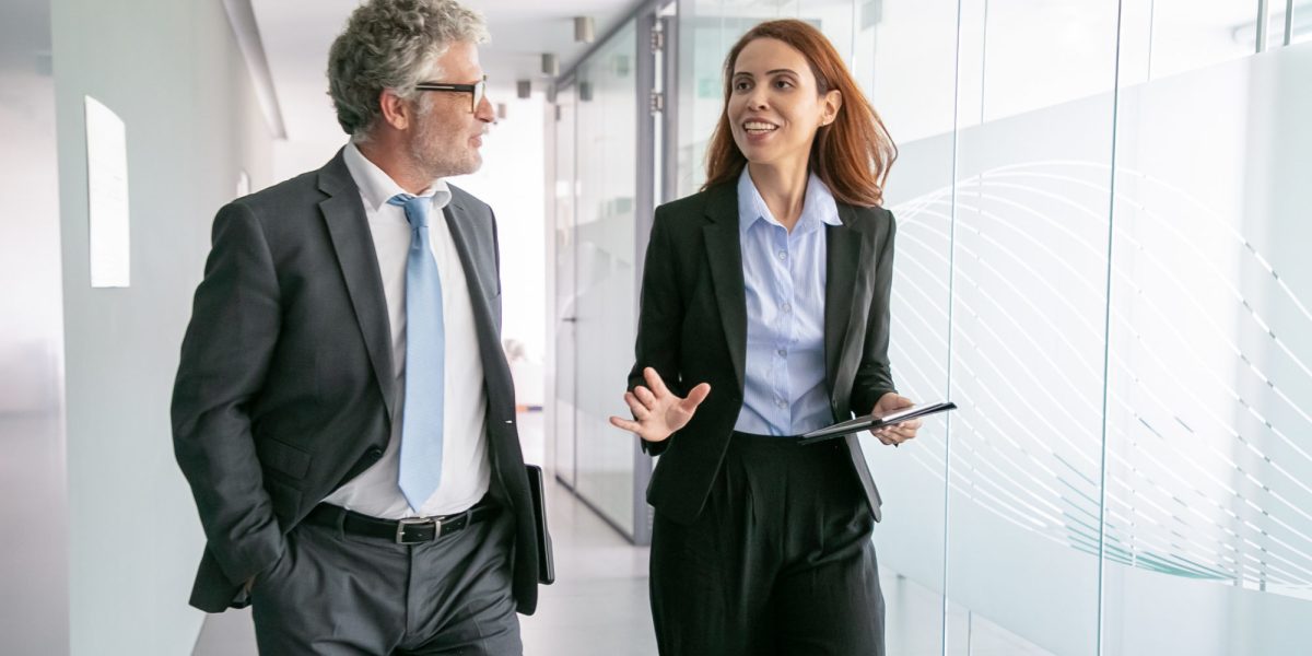 Successful businesspeople walking through office corridor and talking. Professional senior executive manager in eyeglasses listening beautiful content businesswoman. Business and management concept